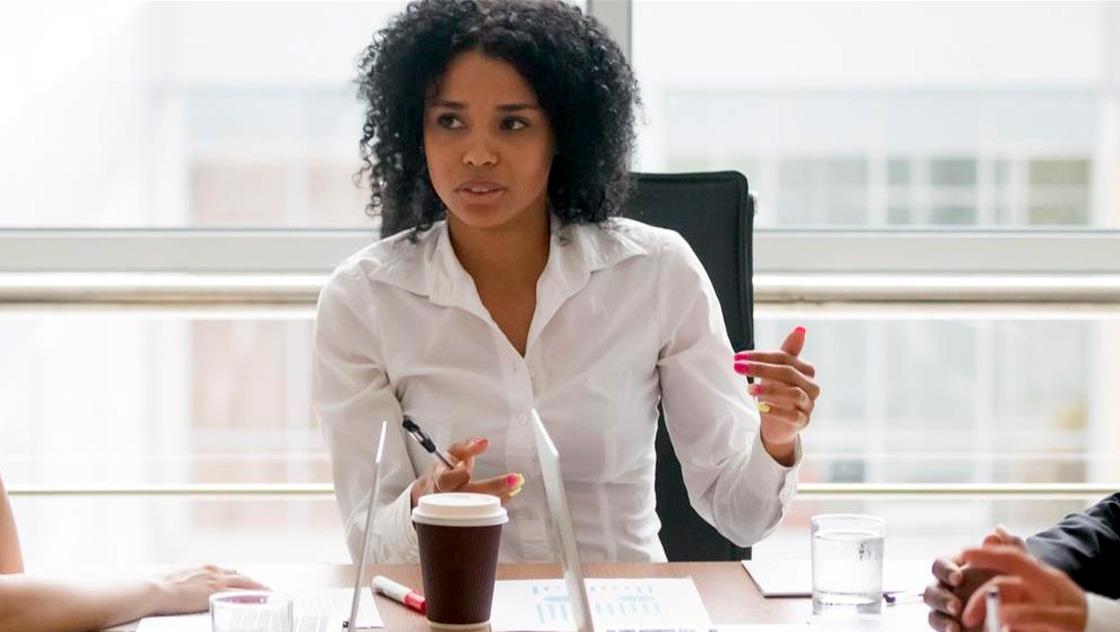 A woman speaks to two colleagues during a meeting, engaged in discussion around a table.