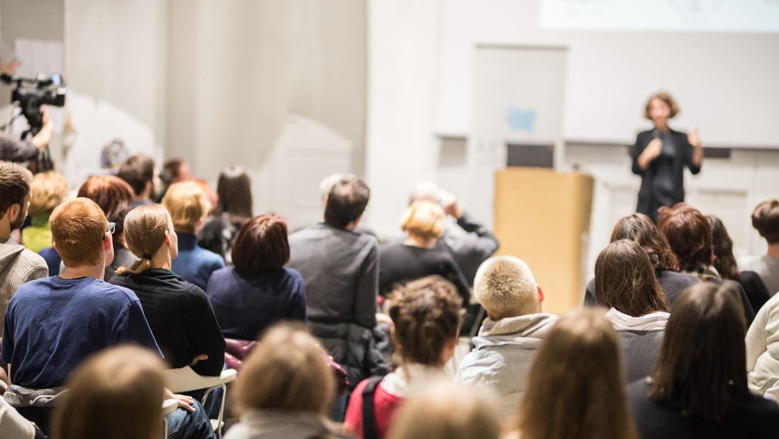 Audience watching a speaker at a conference, with a camera recording the event.