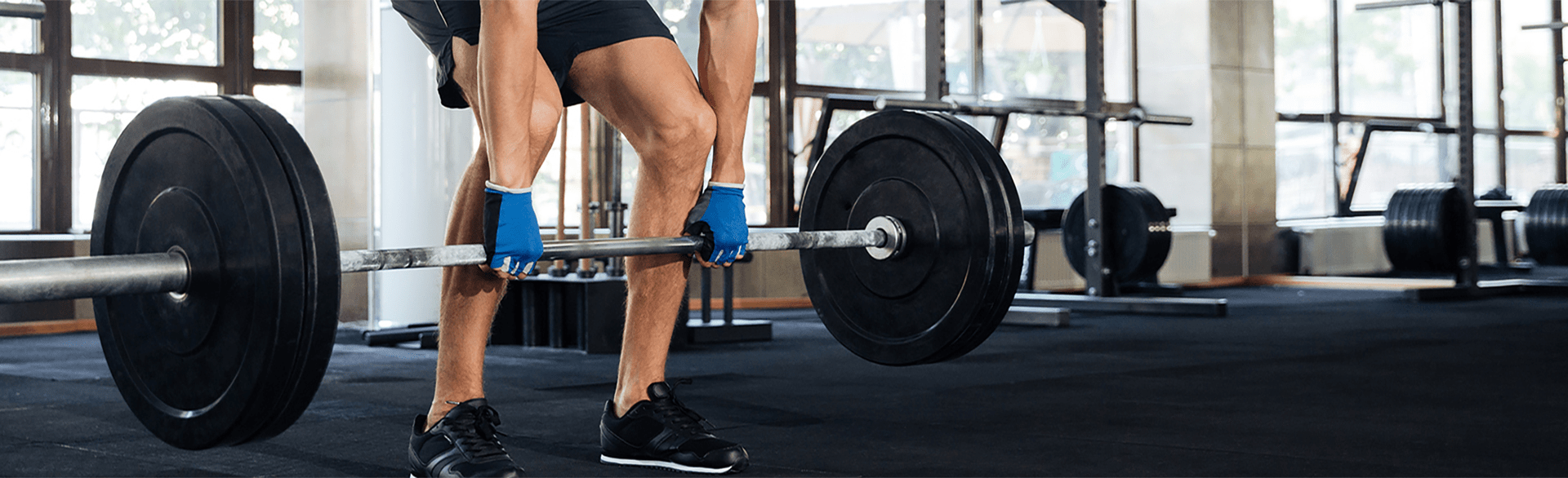 Person lifting a barbell in a gym with large windows and equipment in the background.