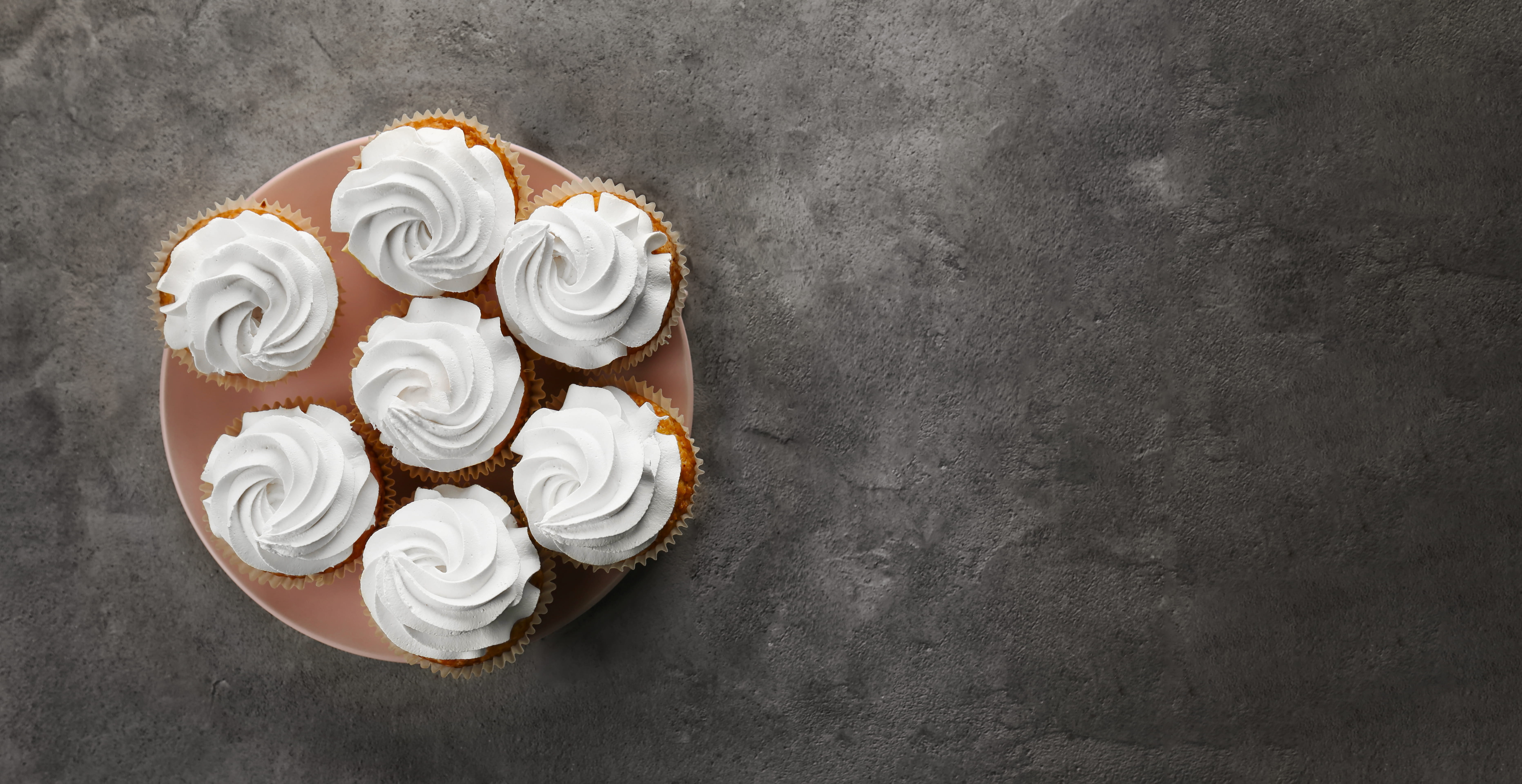 Seven frosted cupcakes on pink plate over dark background.