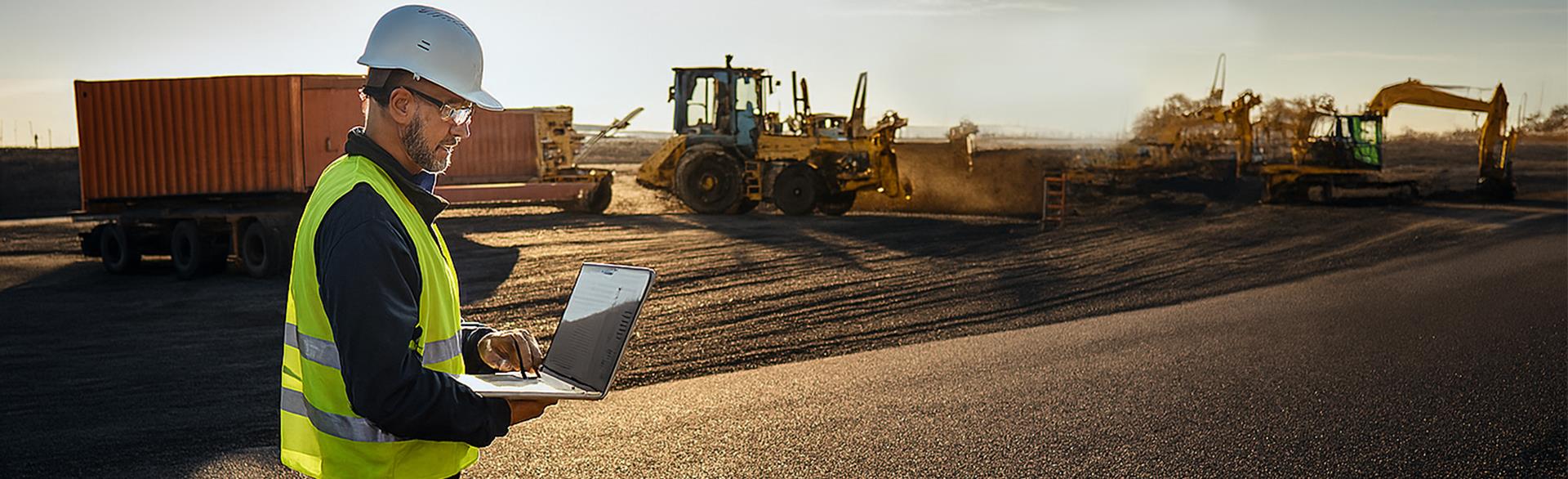 Construction worker with laptop at site with heavy machinery.