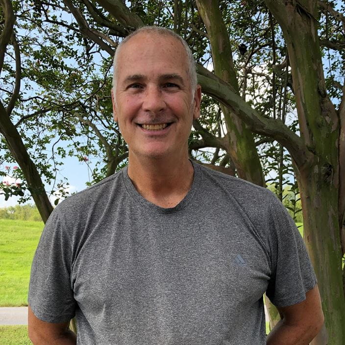 Person standing outdoors in front of a tree, wearing a gray t-shirt.