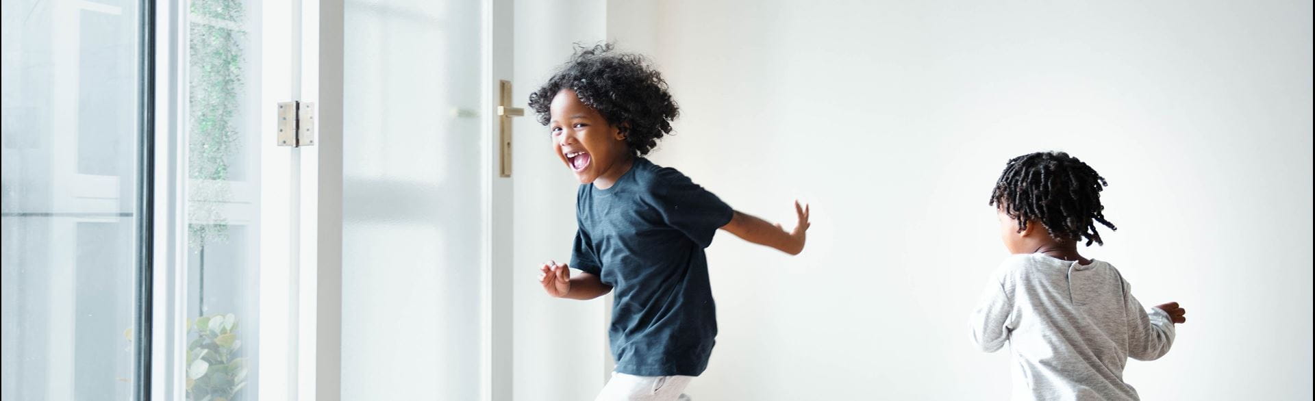Two children running indoors on wooden flooring near large glass doors.