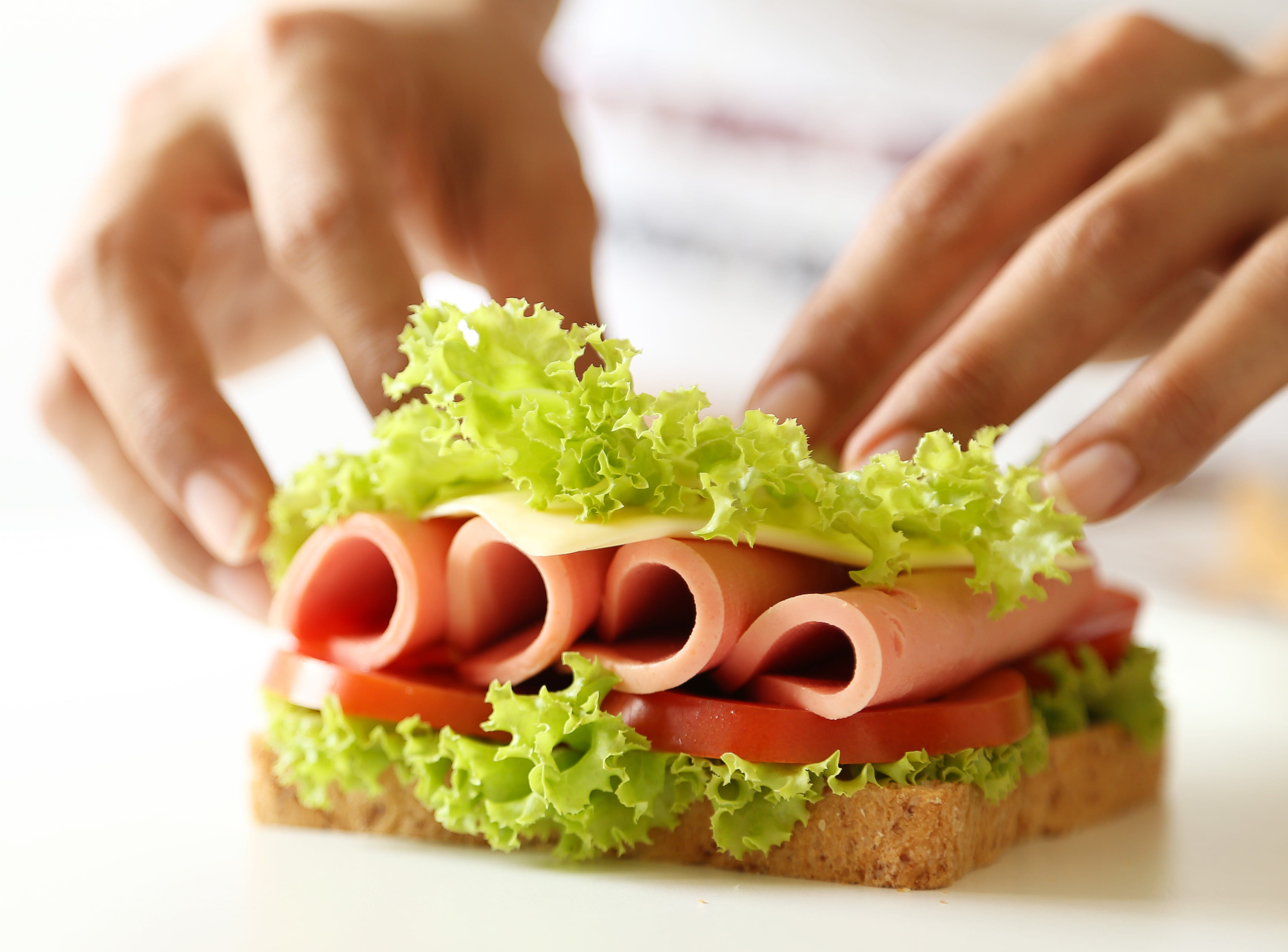 Sandwich being assembled with lettuce, tomato, deli meat, and cheese, held by two hands