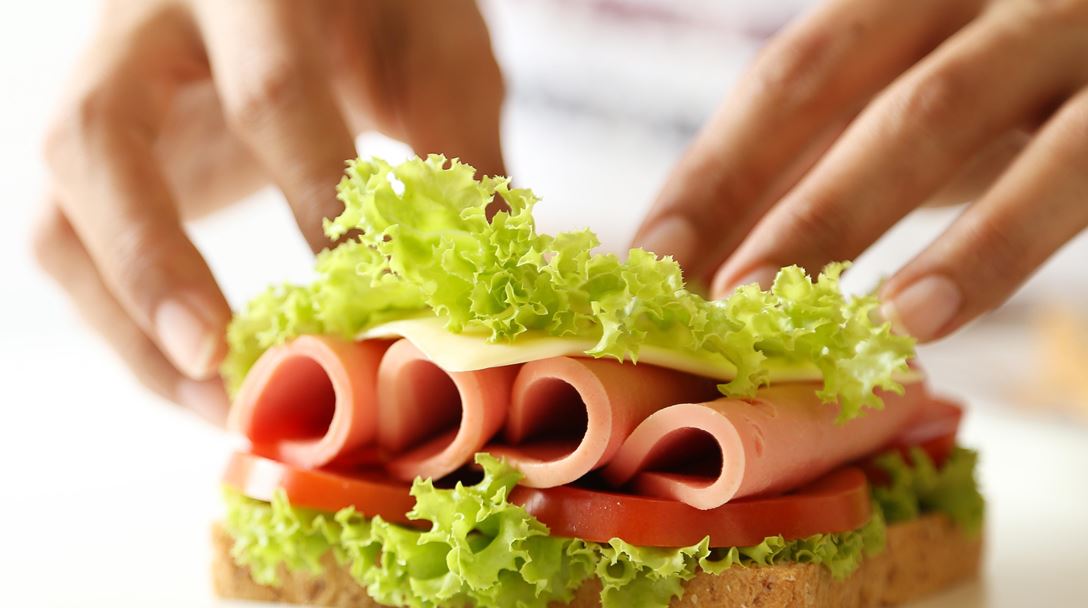 Sandwich being assembled with lettuce, tomato, deli meat, and cheese, held by two hands