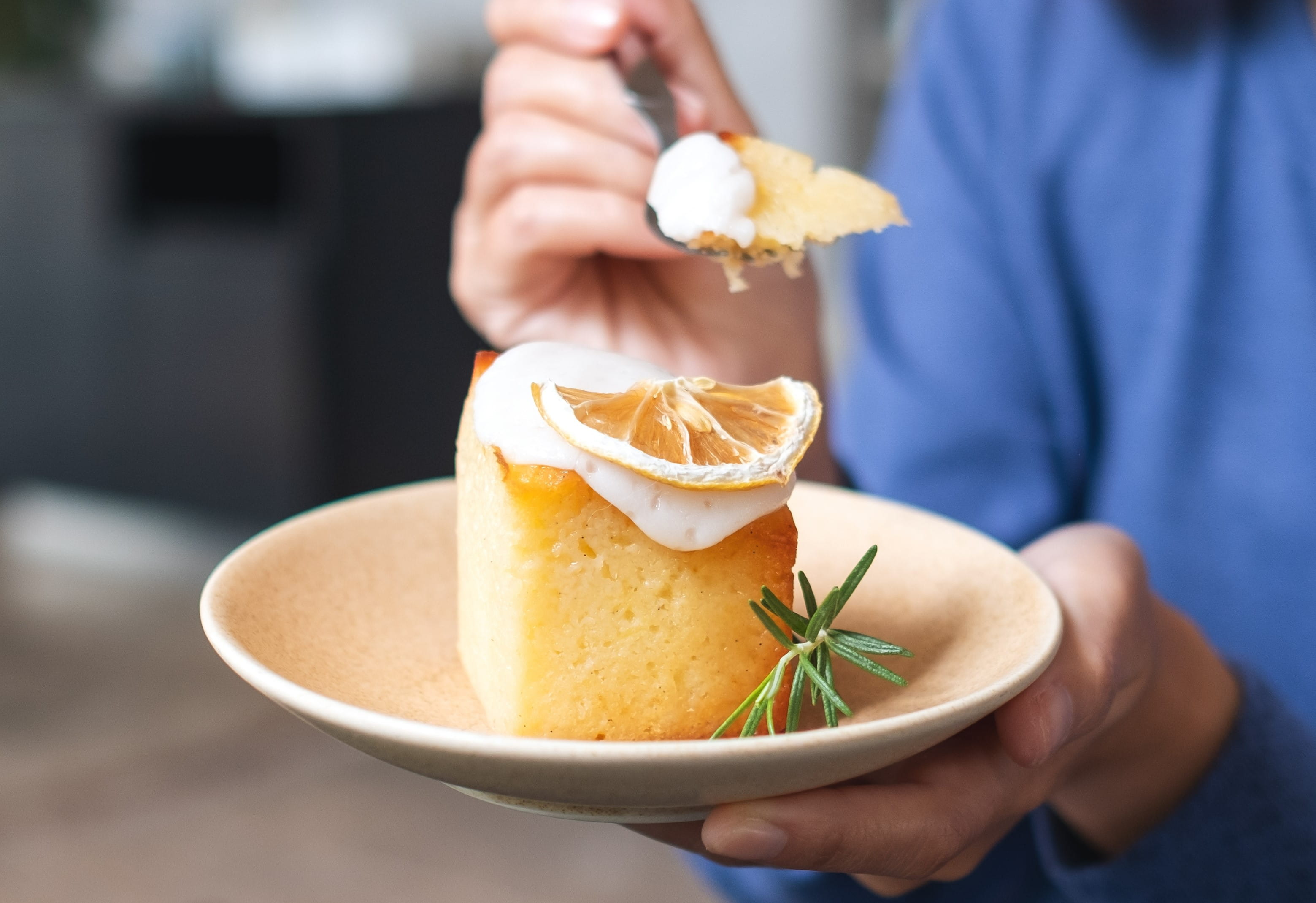 Person holding a plate with lemon-topped cake, whipped cream, and rosemary, taking a bite with a fork.
