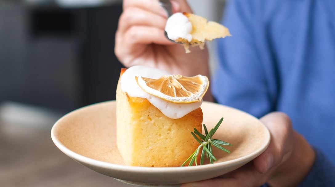 Person holding a plate with lemon-topped cake, whipped cream, and rosemary, taking a bite with a fork.