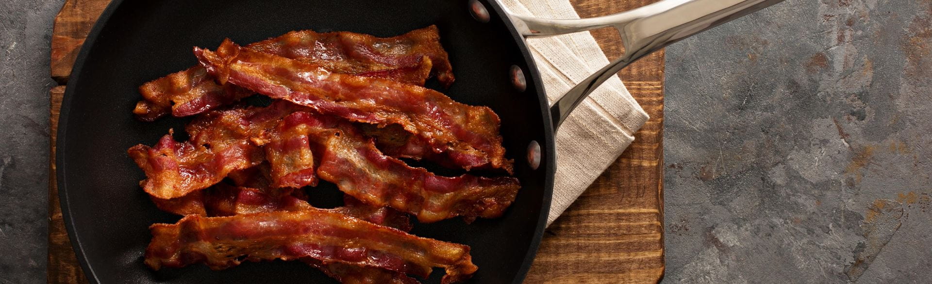 Black frying pan with cooked bacon strips on a wooden cutting board, set on a gray countertop.