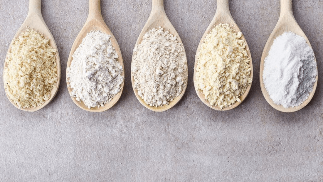 Five wooden spoons with different flours on a gray surface