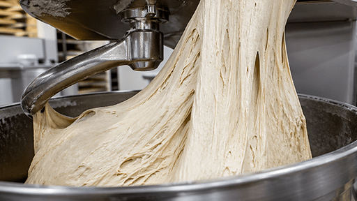 Industrial mixer stretching elastic dough in metal bowl.