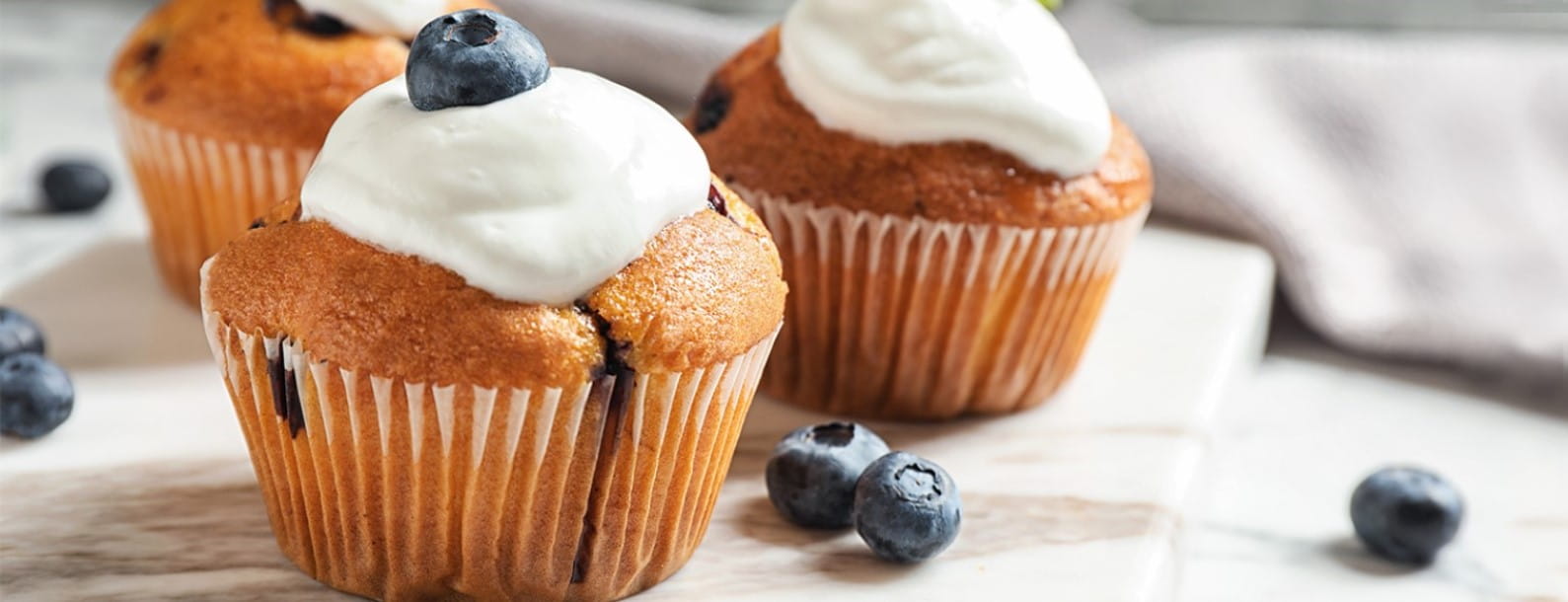 Three blueberry muffins with cream and blueberries, surrounded by loose berries