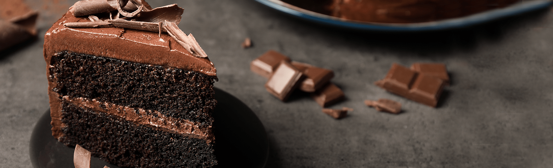 Slice of chocolate cake with shavings on a black plate, surrounded by chocolate pieces