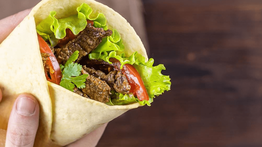 Hand holding a tortilla wrap filled with lettuce, grilled meat, tomatoes, and cilantro