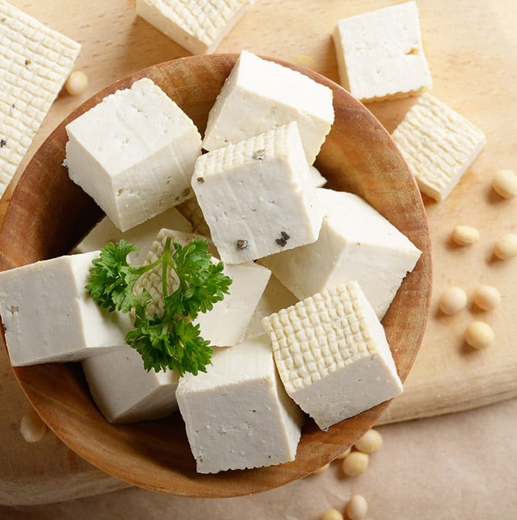 Wooden bowl of tofu cubes with parsley garnish and soybeans on a wooden surface