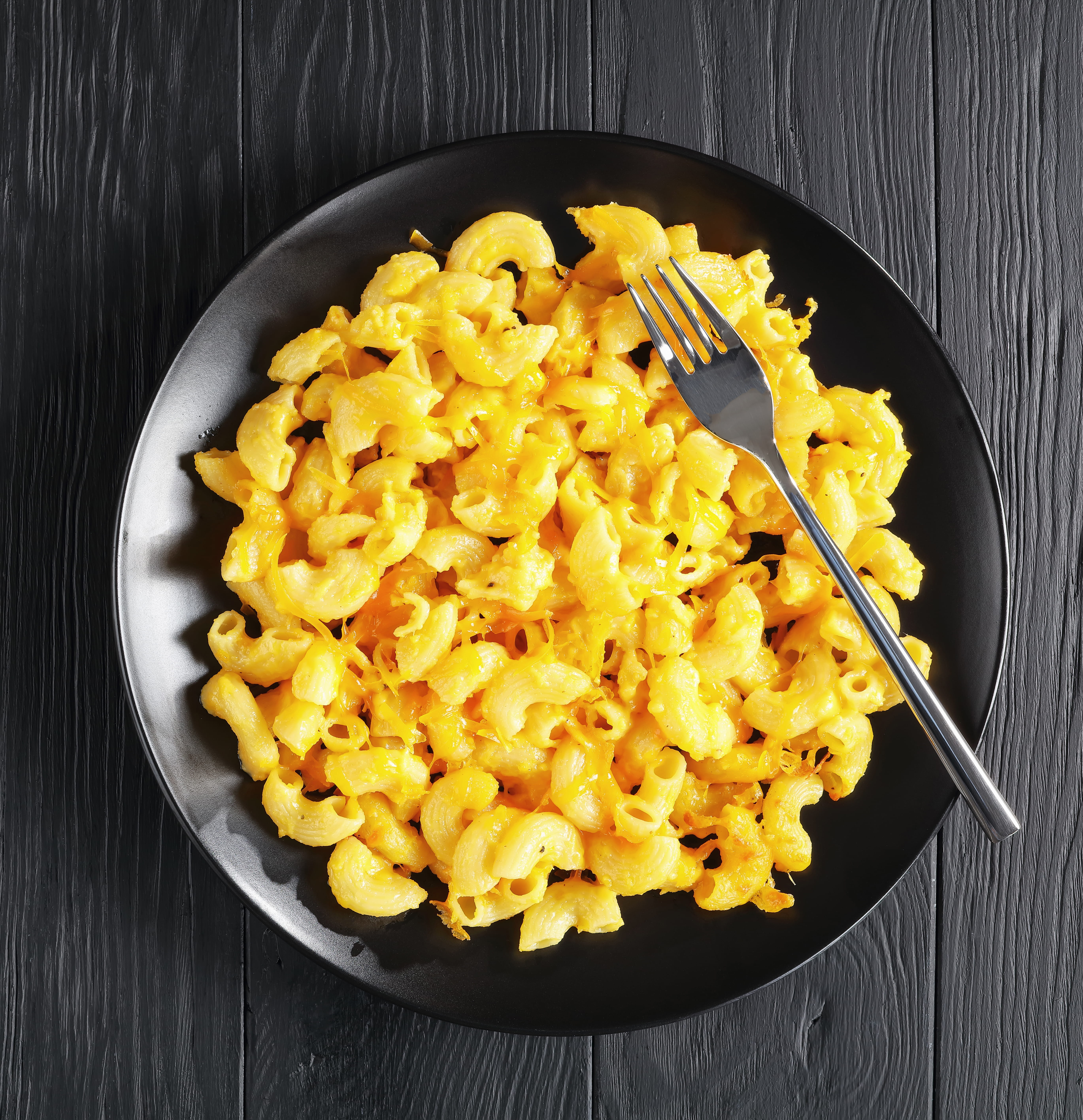 Black plate of macaroni and cheese with a fork on a dark wooden surface