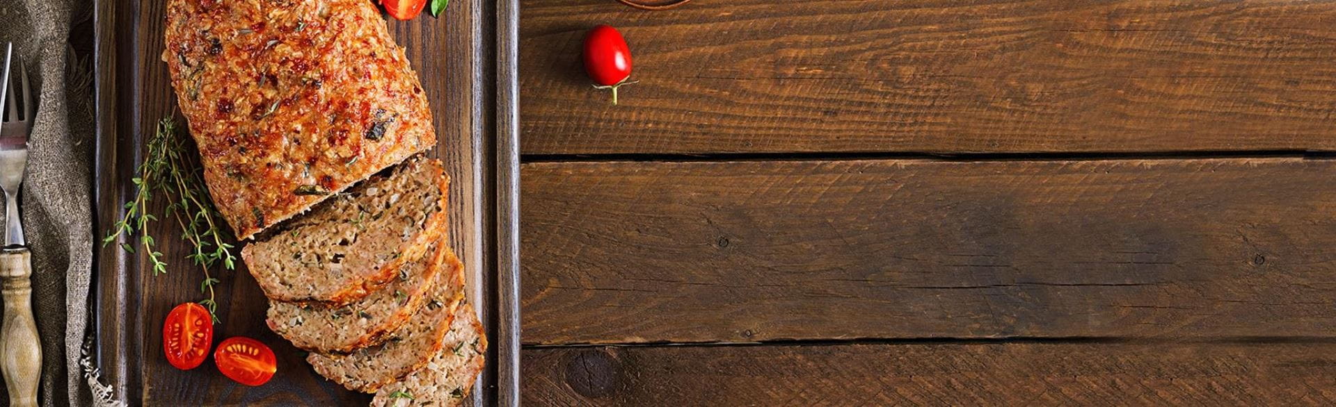 Partially sliced loaf of bread on a cutting board with cherry tomatoes, herbs, fork, and knife