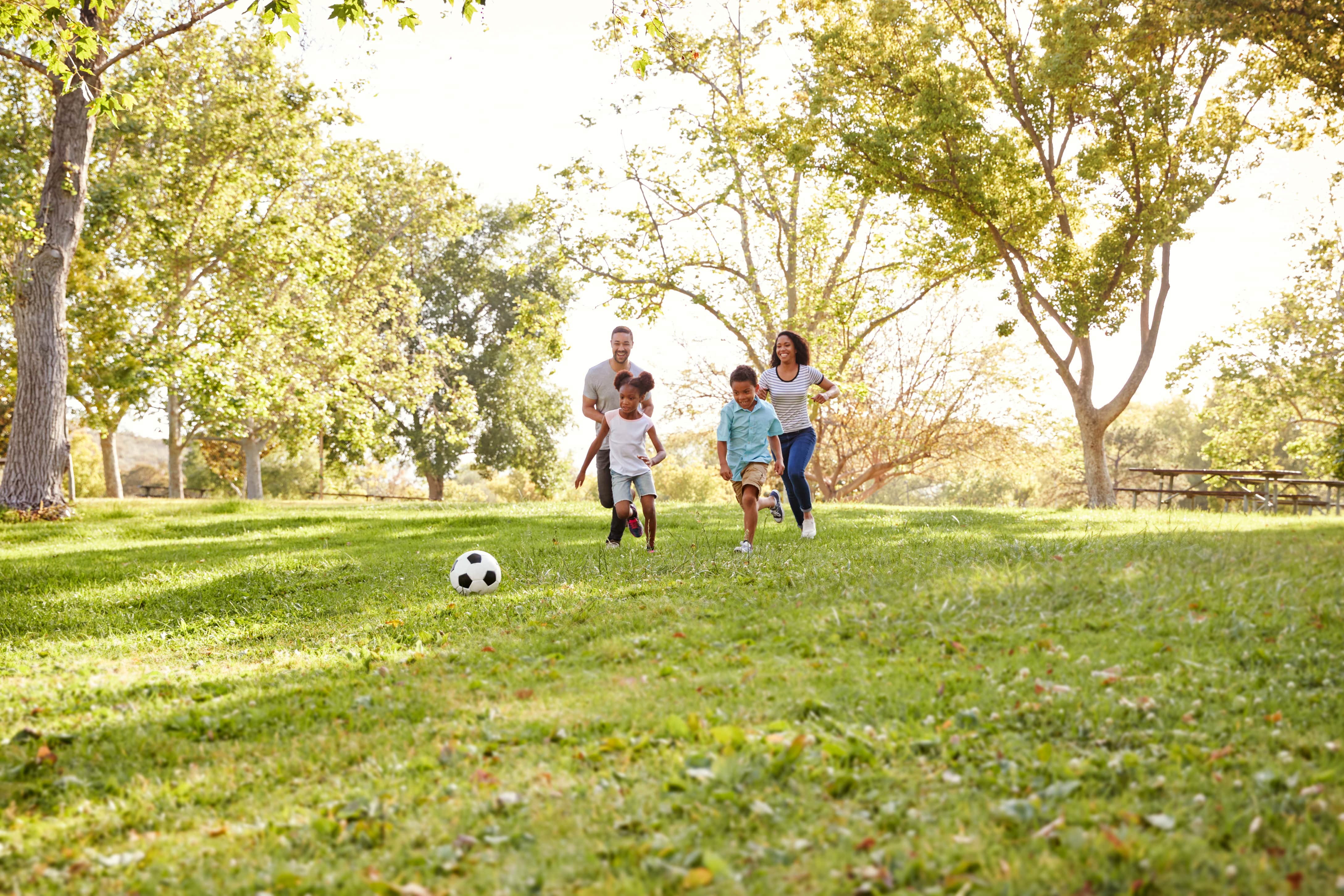 Family of four running in a grassy park with a soccer ball and trees in the background