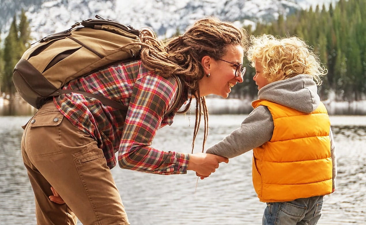 Adult and child holding hands near a lake with mountains and trees in the background