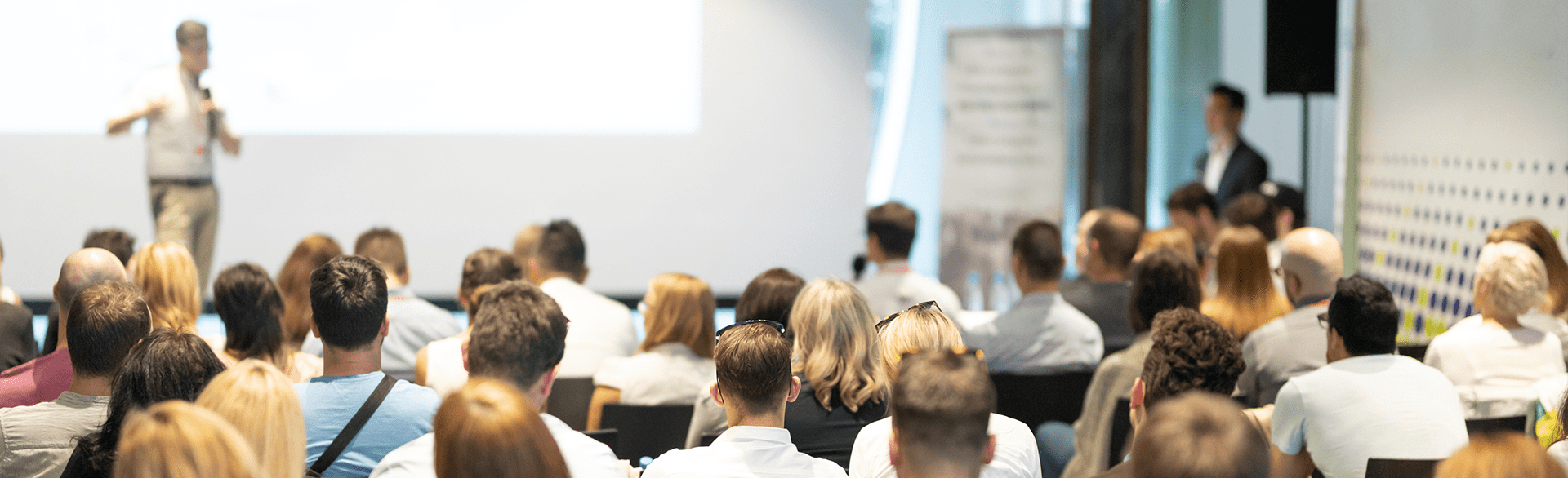 People seated in a conference room listening to a speaker in front of a screen.
